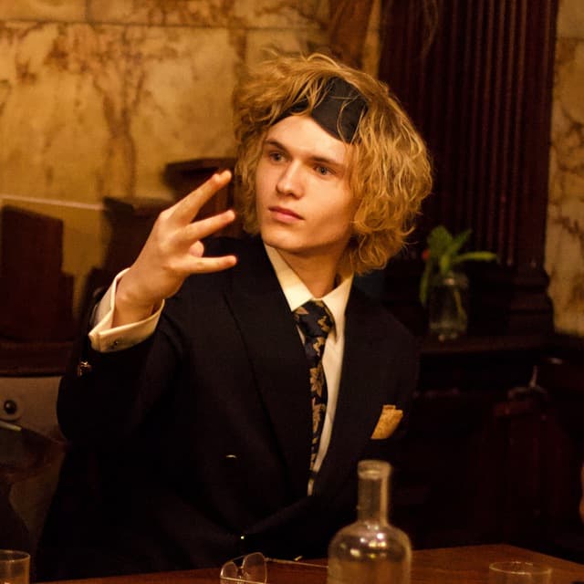 Sam Human in a dark suit and patterned tie, sitting at a table in a warmly lit bar and gesturing while talking