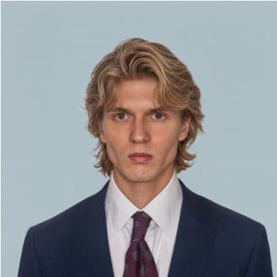 Sam Human, head and shoulders, in a navy suit, white shirt and red tie against a pale blue background