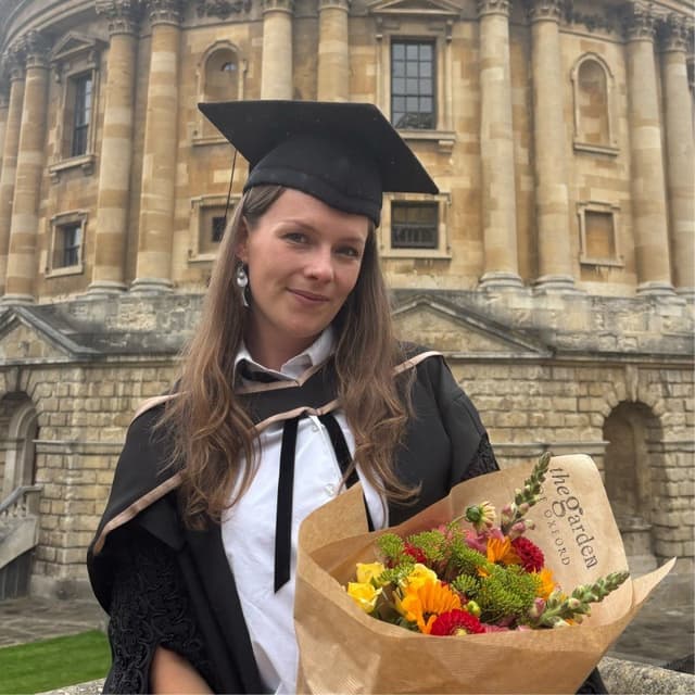 Sophie Kitchen in a graduation gown and cap, holding a bouquet of flowers, in front of a domed stone building